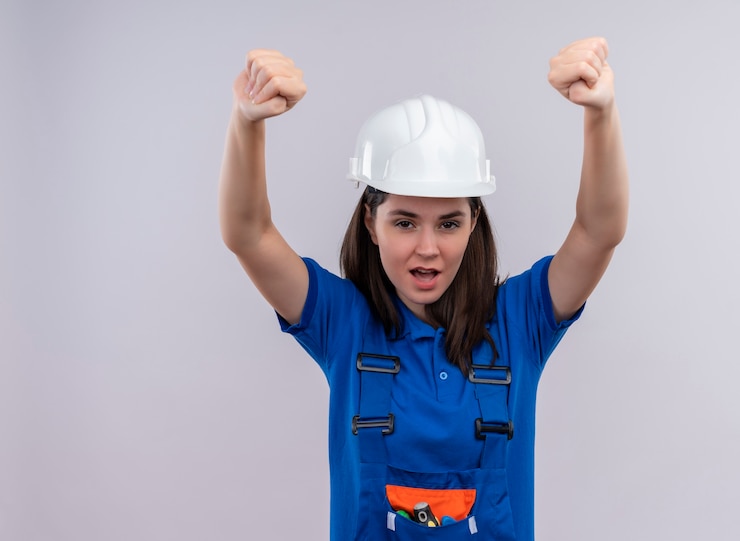 Foto grátis construtora jovem confiante com capacete de segurança branco e uniforme azul levanta os punhos e olha para a câmera em um fundo branco isolado com espaço de cópia