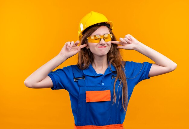 Construtora jovem trabalhadora em uniforme de construção e capacete de segurança fechando as orelhas com os dedos para o barulho do som alto em pé sobre a parede laranja