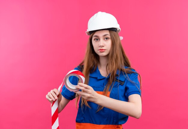 Construtora jovem trabalhadora em uniforme de construção e capacete de segurança segurando fita adesiva e olhando com cara séria em pé sobre a parede rosa