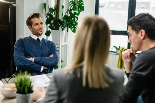 Consultor financeiro sorridente sentado com os braços cruzados e conversando com um casal sobre seus futuros planos de investimento