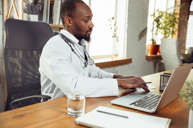 Consultoria médica para paciente, trabalhando com laptop. Médico afro-americano durante seu trabalho com pacientes, explicando receitas de remédios. Trabalho árduo diário pela saúde e salvamento de vidas durante a epidemia.