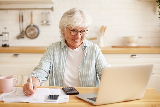 Contadora aposentada alegre trabalhando distante de casa usando um computador portátil genérico, sentada à mesa da cozinha com calculadora e telefone celular, segurando um lápis, fazendo anotações em documentos financeiros