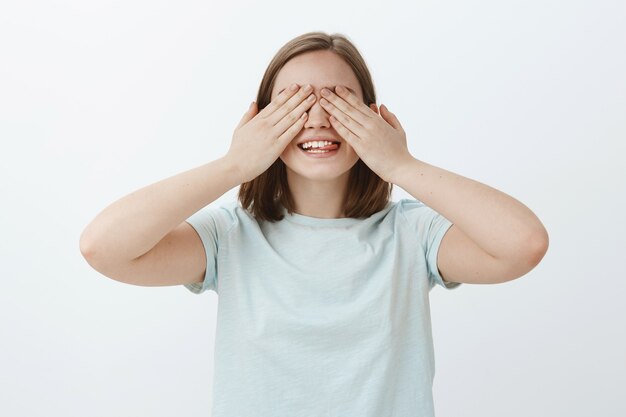 Contando dez e indo te procurar. Retrato de uma mulher divertida e emotiva esperando a surpresa com as palmas das mãos nos olhos, sorrindo alegremente de empolgação enquanto amigos trazem um presente secreto sobre a parede cinza