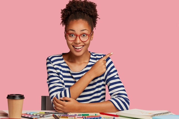 Foto grátis contente artesã negra em roupas listradas, mostra espaço livre contra parede rosa, trabalha em novo esboço em caderno com giz de cera, bebe leva café, trabalha em casa, tem habilidades criativas