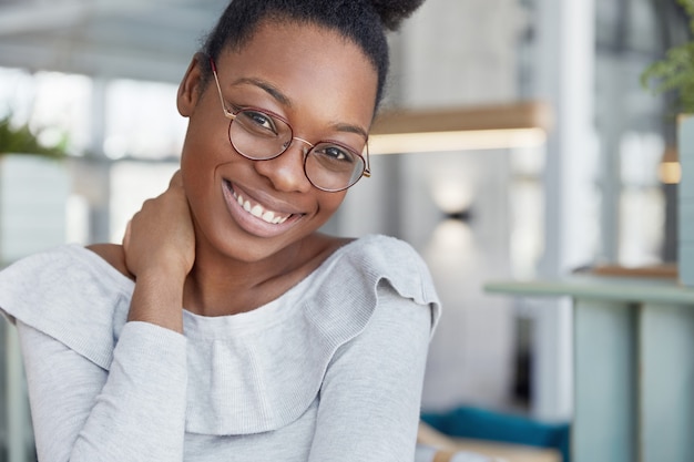 Contente e atraente modelo feminino de pele escura usa óculos, tem um sorriso brilhante, está feliz por terminar o trabalho e tem pausa, posa contra o interior do escritório.