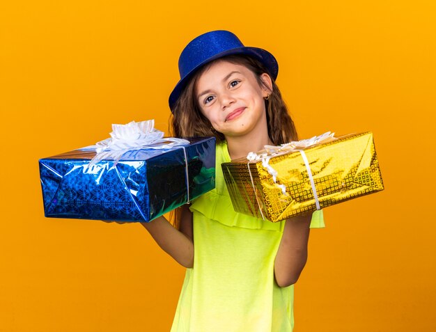 contente garotinha caucasiana com chapéu de festa azul segurando caixas de presente isoladas na parede laranja com espaço de cópia