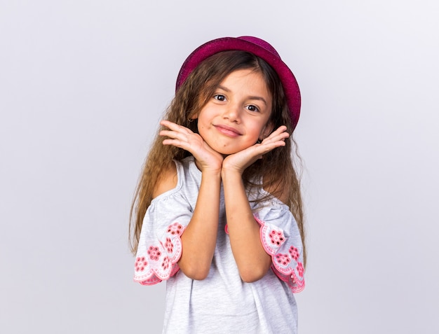 Contente garotinha caucasiana com chapéu de festa roxo colocando as mãos no queixo isolado na parede branca com espaço de cópia