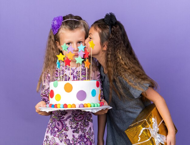 contente garotinha caucasiana segurando uma caixa de presente e beijando a triste garotinha loira segurando um bolo de aniversário isolado na parede roxa com espaço de cópia