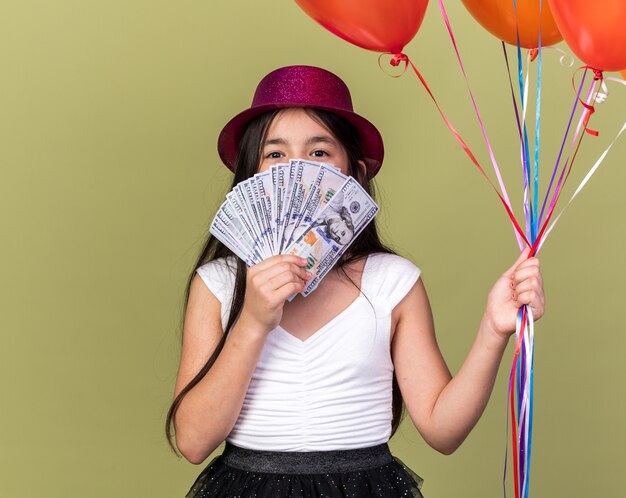 contente jovem caucasiana com chapéu de festa roxo segurando balões de hélio e dinheiro na frente do rosto, isolado na parede verde oliva com espaço de cópia