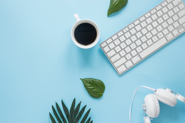 Copo de café branco; fone de ouvido e teclado com folhas no pano de fundo azul