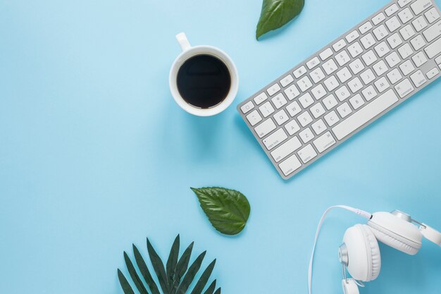 Copo de café branco; fone de ouvido e teclado com folhas no pano de fundo azul