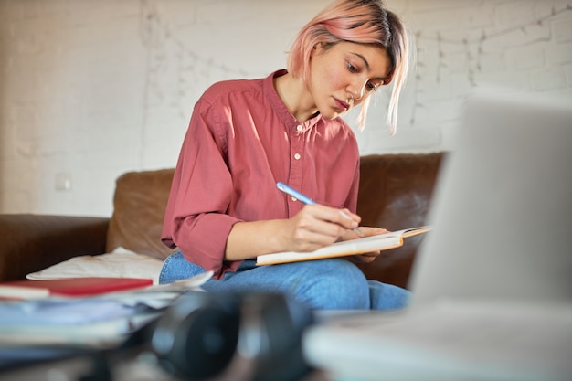 Copywriter jovem concentrado com cabelo rosa, trabalhando em casa, fazendo anotações no caderno.