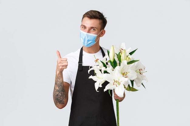 Coronavirus, distanciamento social dos negócios durante o conceito de pandemia covid-19. florista simpática e simpática, vendedor em exposição de flores, mostrando o polegar para cima, sorrindo e segurando buquê de lírios, usa máscara médica