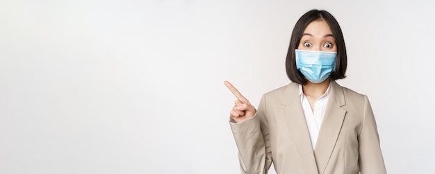 Coronavirus e conceito de trabalho retrato de mulher com máscara facial médica apontando o dedo para a esquerda mostrando o logotipo ou o banner de fundo branco