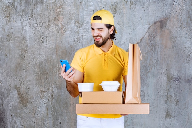 Correio com uniforme amarelo segurando pacotes para viagem e sacola de compras e fazendo uma videochamada