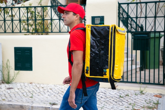 Correio concentrado entregando pedidos e andando pela rua. Entregador profissional carregando mochila amarela e procurando o endereço necessário. Serviço de entrega e conceito de compras online