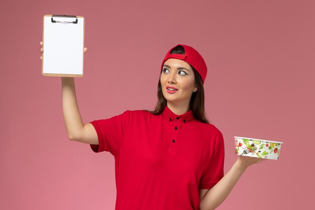 Foto grátis correio feminino com capa de uniforme vermelho e bloco de notas de tigela redonda nas mãos na parede rosa claro, trabalho de funcionário de entrega de uniforme.