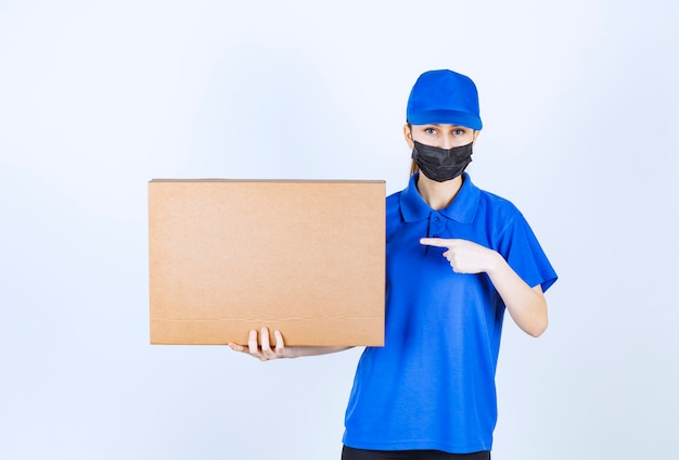 Correio feminino com máscara e uniforme azul, segurando um grande pacote de papelão.