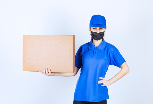 Correio feminino com máscara e uniforme azul, segurando um grande pacote de papelão.