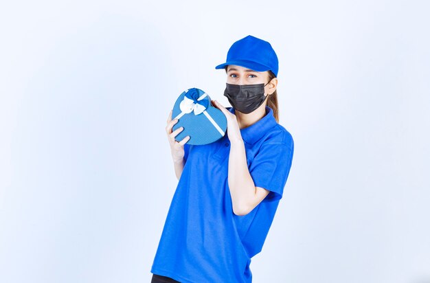 Correio feminino com máscara e uniforme azul, segurando uma caixa de presente em forma de coração.