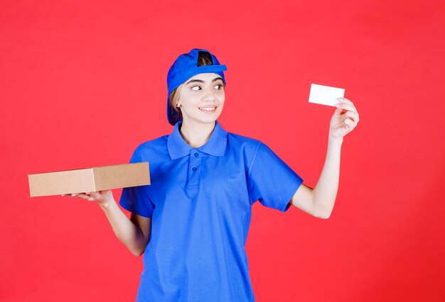 Correio feminino com uniforme azul, segurando uma caixa para viagem e apresentando seu cartão de visita.