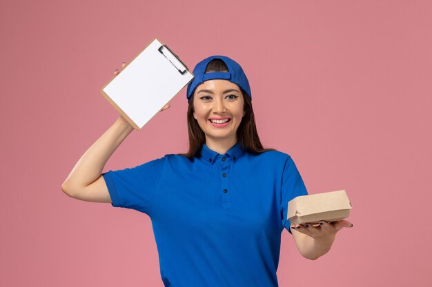 Correio feminino de frente para a capa uniforme azul segurando um pequeno pacote de entrega vazio com o bloco de notas na parede rosa, trabalhador de entrega de empresa de serviço de funcionário