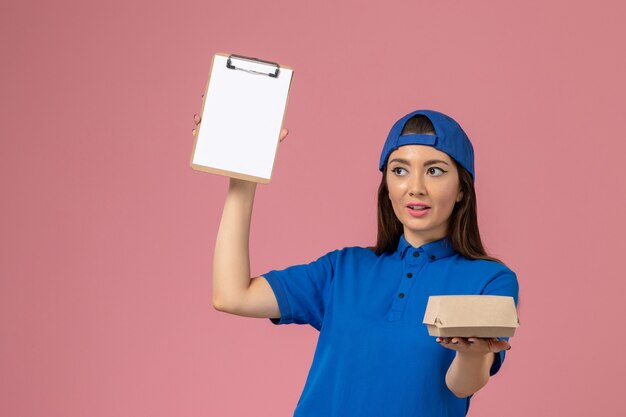 Correio feminino de frente para a capa uniforme azul segurando um pequeno pacote de entrega vazio com o bloco de notas na parede rosa, trabalho de entrega de empresa de serviço de funcionário