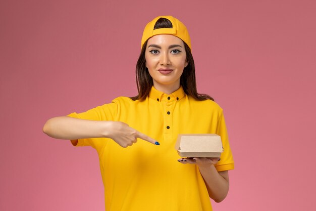 Correio feminino de frente para o uniforme amarelo e capa segurando um pequeno pacote de entrega de comida na parede rosa.