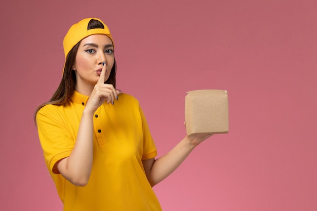 Correio feminino de frente para o uniforme amarelo e capa segurando um pequeno pacote de entrega de comida no uniforme rosa claro serviço de entrega de serviço de empresa de trabalho