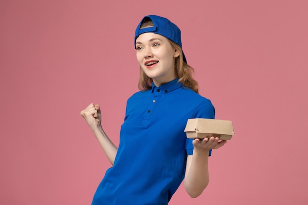 Correio feminino de frente para o uniforme azul e capa segurando um pequeno pacote de entrega de comida no fundo rosa uniforme de entrega de trabalho serviço de empresa trabalhador