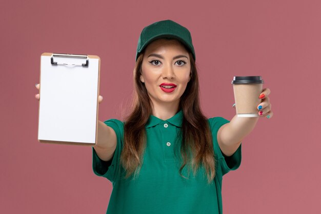 Foto grátis correio feminino de frente para o uniforme verde e capa segurando a xícara de café de entrega e o bloco de notas na parede rosa.