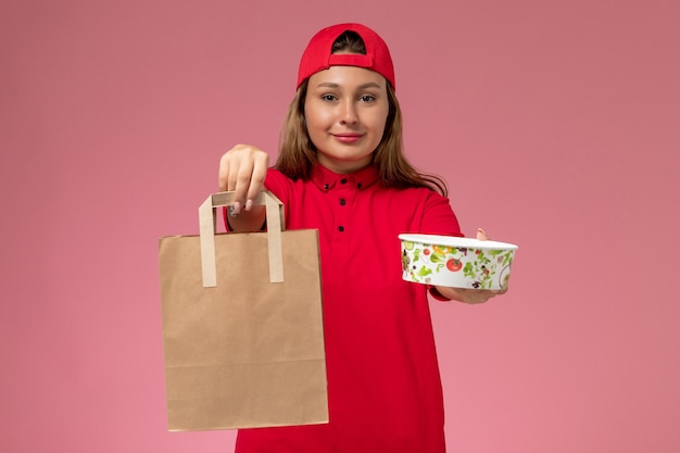 Correio feminino de frente para o uniforme vermelho e capa segurando o pacote de comida de entrega e a tigela na parede rosa claro, serviço de entrega uniforme