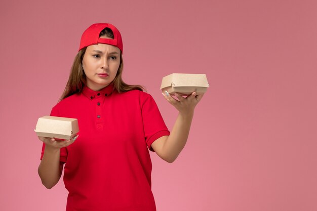 Correio feminino de frente para o uniforme vermelho e capa segurando pacotes de comida de entrega e pensando na parede rosa claro, empresa de trabalhador de trabalho de serviço de entrega de uniforme