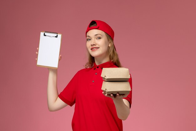 Correio feminino de frente para o uniforme vermelho e capa segurando pequenos pacotes de comida de entrega e bloco de notas na parede rosa claro, trabalho de entrega de uniforme de serviço