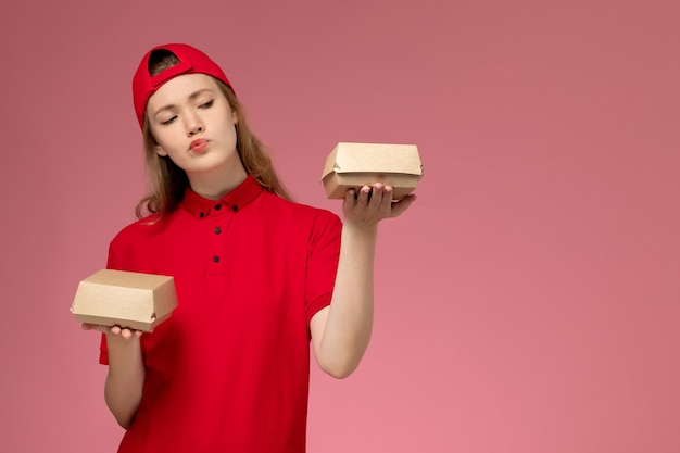 Correio feminino de frente para o uniforme vermelho e capa segurando pequenos pacotes de comida de entrega na parede rosa, trabalho de uniforme de empresa de serviço de entrega de trabalho