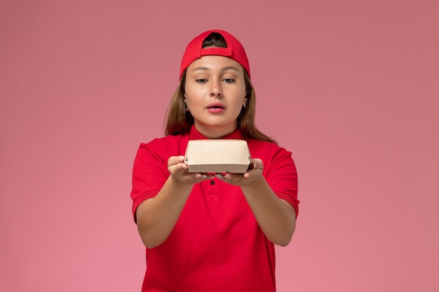 Correio feminino de frente para o uniforme vermelho e capa segurando um pequeno pacote de entrega de comida na parede rosa claro, trabalho de empresa de serviço de entrega de uniforme