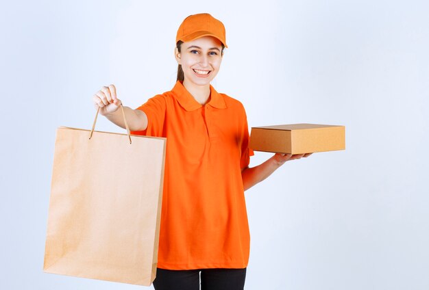 Correio feminino de uniforme amarelo entregando uma sacola de compras e uma caixa de papelão ao cliente.