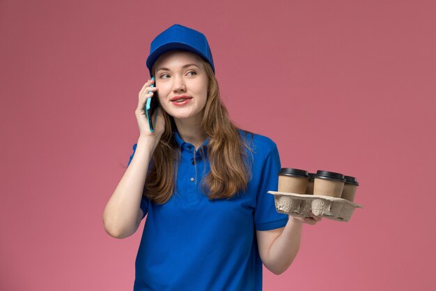 Correio feminino de uniforme azul, de vista frontal, falando ao telefone segurando xícaras de café de entrega na empresa de uniforme de serviço de mesa de luz rosa