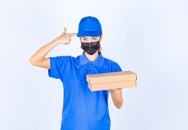 Correio feminino de uniforme azul e máscara facial, segurando uma caixa de papelão e pensando na entrega.