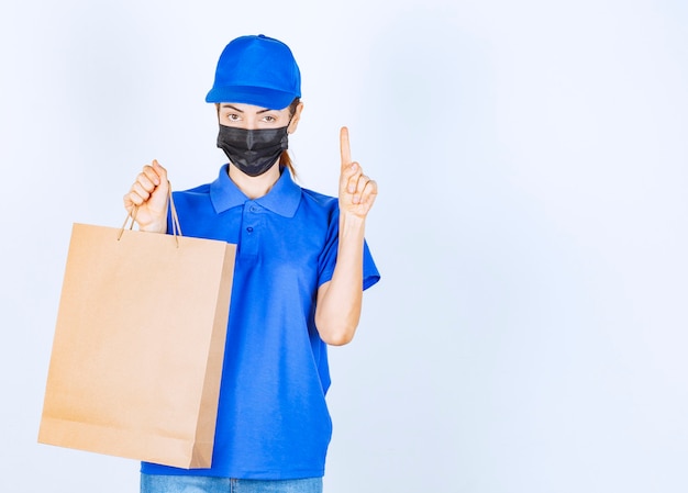 Correio feminino de uniforme azul e máscara facial, segurando uma sacola de compras de papelão e tem uma ideia ou pensamento.