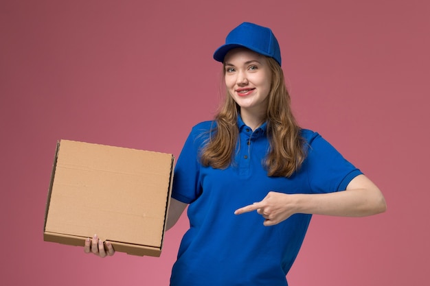 Correio feminino de uniforme azul segurando uma caixa de comida com um sorriso na mesa rosa.