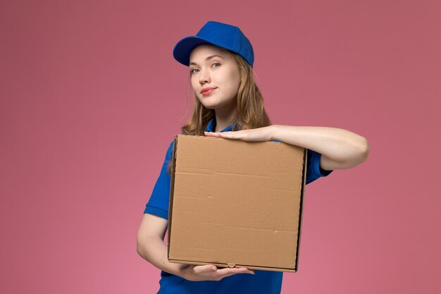 Correio feminino de uniforme azul segurando uma caixa de entrega de comida na mesa rosa.