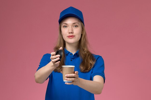 Correio feminino de uniforme azul segurando uma xícara de café marrom de frente e abrindo-o na mesa rosa claro.