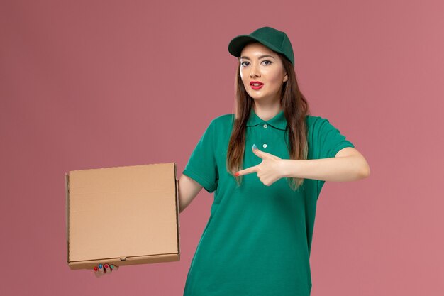 Correio feminino de uniforme verde segurando uma caixa de comida na parede rosa.