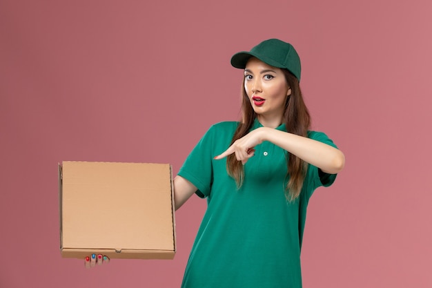 Correio feminino de uniforme verde segurando uma caixa de comida na parede rosa.
