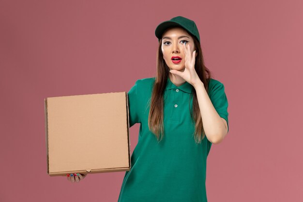 Correio feminino de uniforme verde segurando uma caixa de comida sussurrando na parede rosa entrega uniforme de serviço