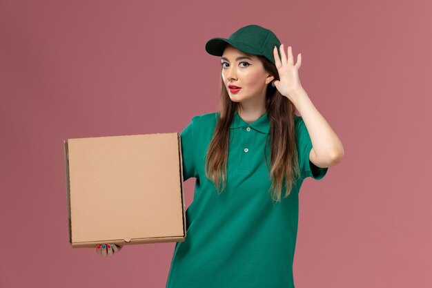 Correio feminino de uniforme verde segurando uma caixa de comida tentando ouvir na parede rosa