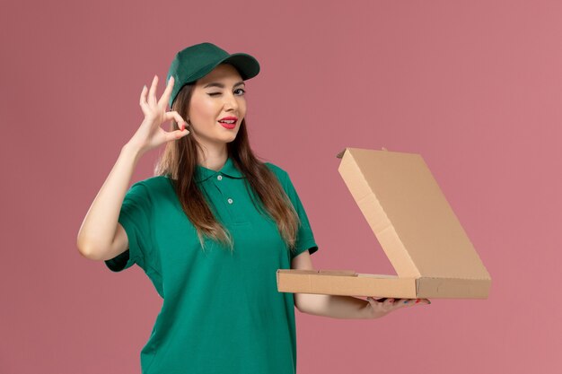 Correio feminino de uniforme verde segurando uma caixa de entrega de comida na parede rosa.