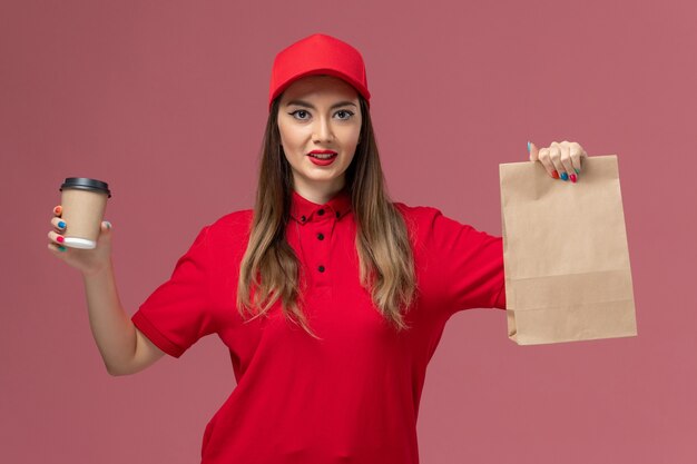 Correio feminino de uniforme vermelho segurando a xícara de café de entrega e o pacote de comida no fundo rosa serviço de entrega uniforme