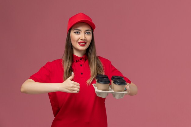 Correio feminino de uniforme vermelho segurando copos de café de entrega posando sorrindo no fundo rosa trabalho de uniforme de entrega